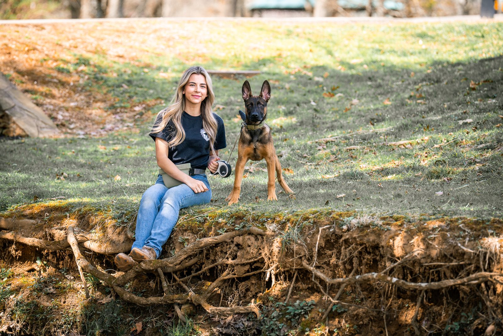 A woman sitting on a mossy bank beside a young Belgian Malinois in warm autumn light